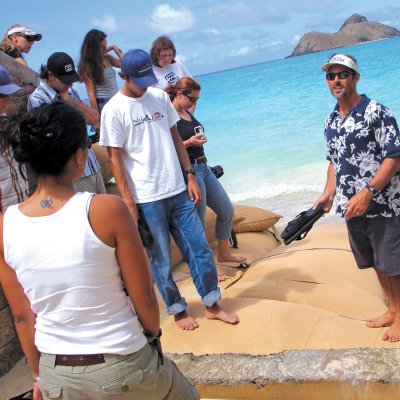 An instructor stands on sandbags on a beach on the east side of Oahu, as students pay close attention