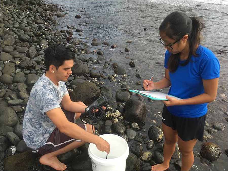 On a rocky shoreline, one student crouches next to a bucket while another stands ready to write on a clipboard
