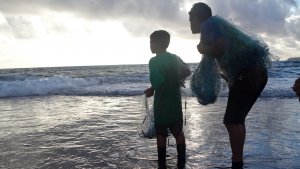 An adult and a child stand on the shoreline and look out into the waves while holding fishing nets about to be cast