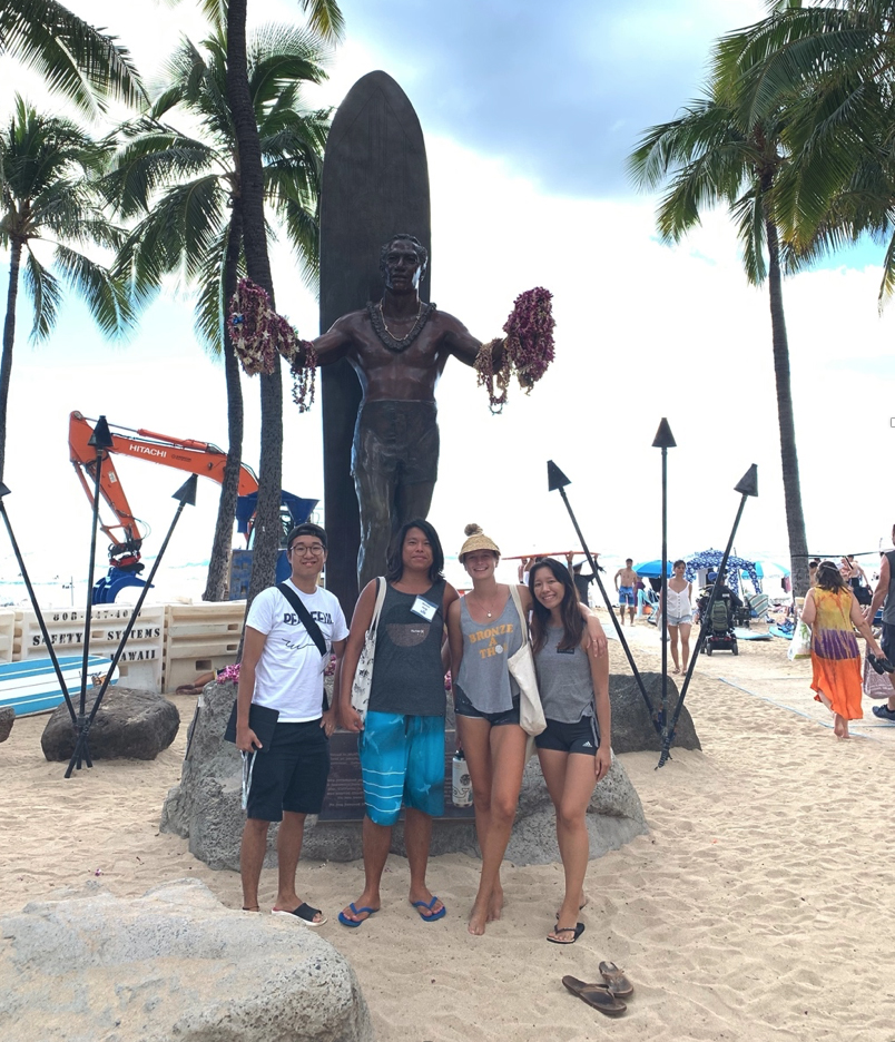 Four students stand in front of a bronze statue with sand underfoot and palm trees behind