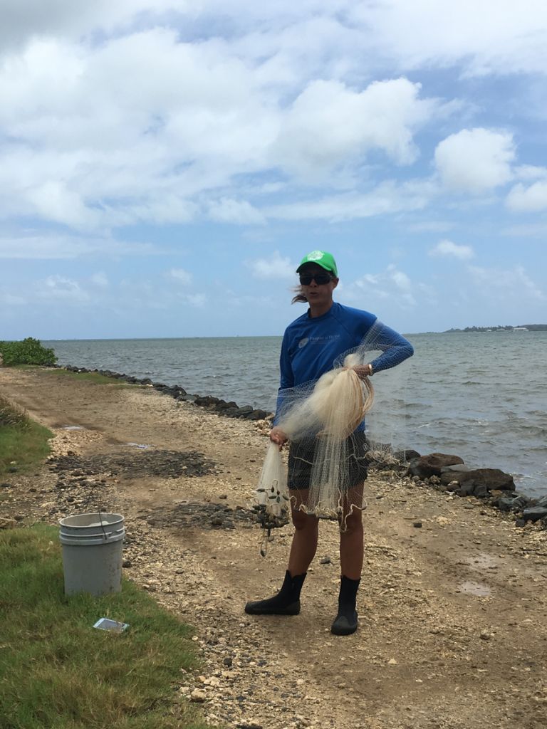 Hi'ilei grips a net while standing on a sandy verge near a low rock wall with open water beyond