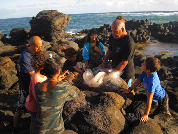 An elder shows students a fishing net at the edge of a rocky pool