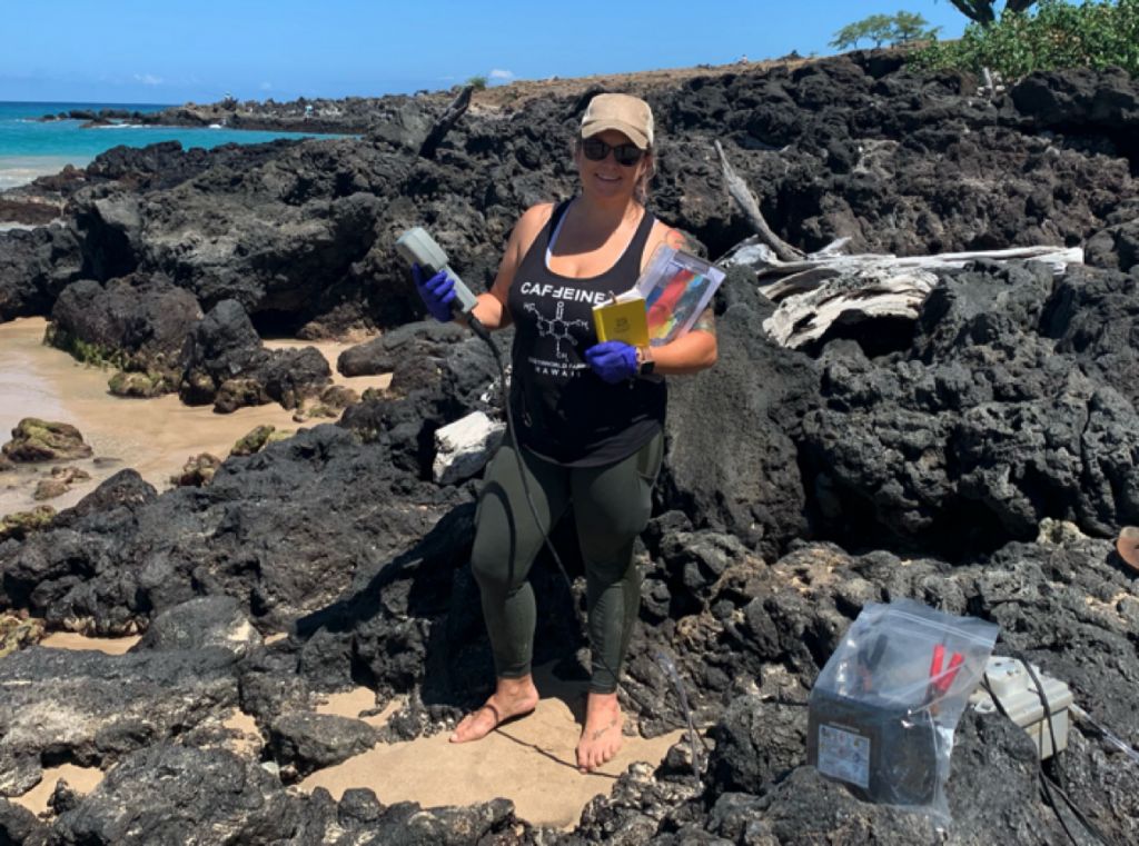 A student stands amidst lava rock and sand with water sampling gear in hand