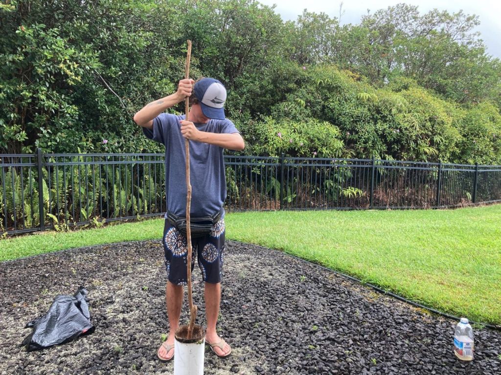 A student stands in a muddy path of a lawn, plunging a long stick into a vertical white tube