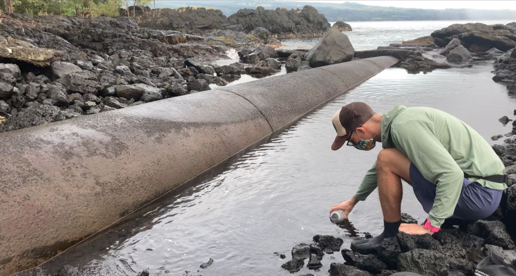 A student crouches to take a water sample from a small water body along a large cement pipe