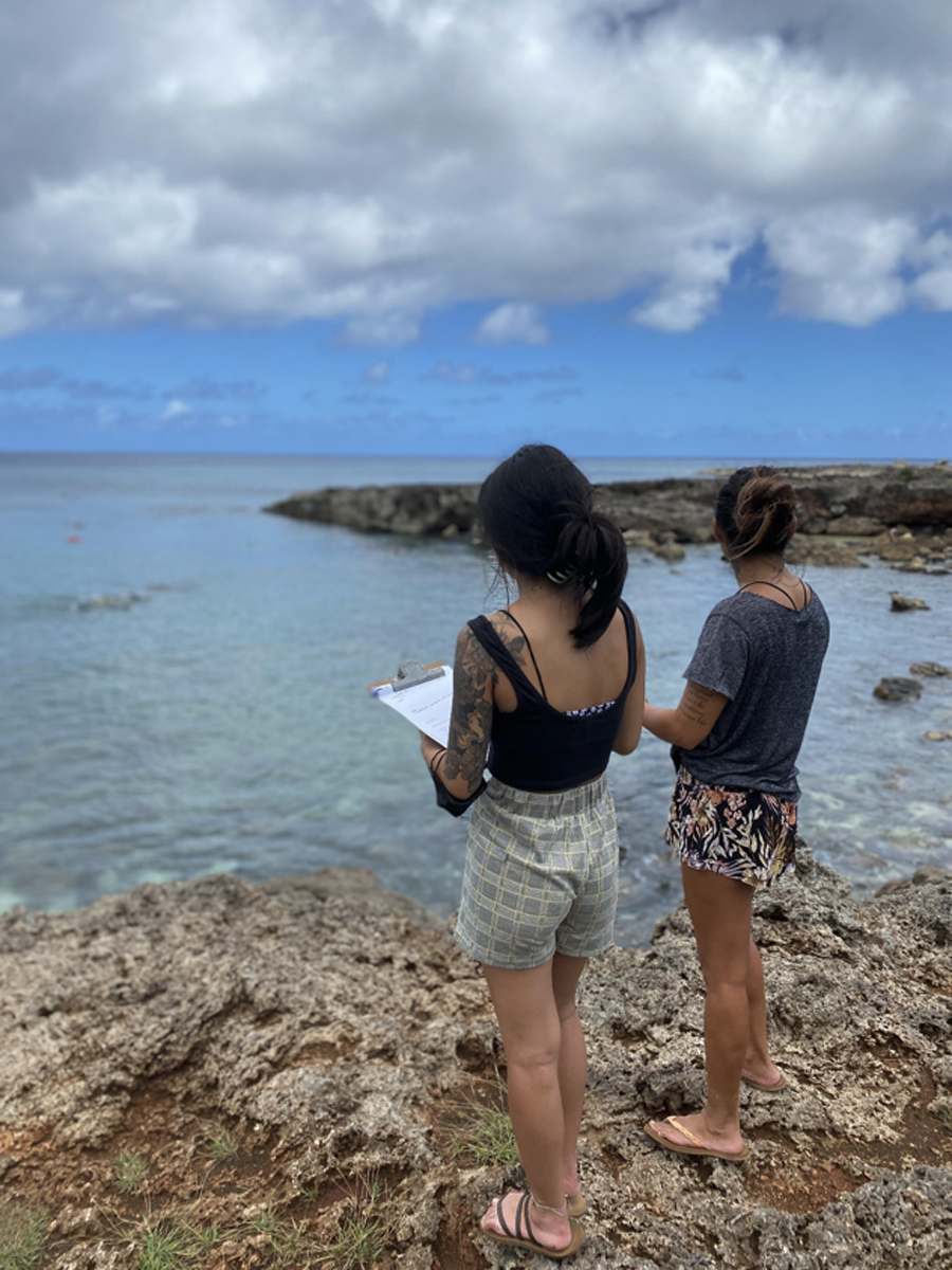 Two young women with clipboards stand on old coral looking out at a rocky inlet