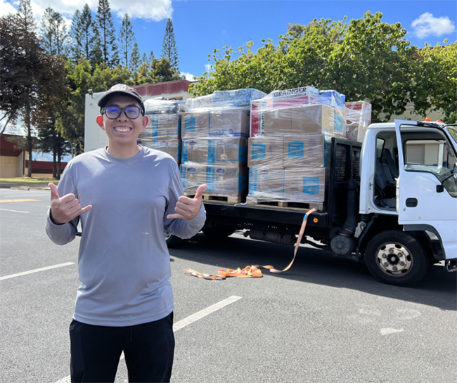 The author poses, grinning, in front of a truck loaded with boxes of supplies