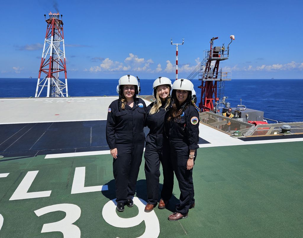 The same three young women pose in flight suits and helmets on the deck of the oil platform with blue ocean and skies behind them