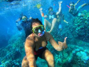 photo of woman snorkeler underwater in a coral reef environment saying hello to the photographer with other snorkelrs behind her.