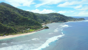 Aerial view of a high, volcanic island with lush greenery and waves meeting a shrinking shoreline.